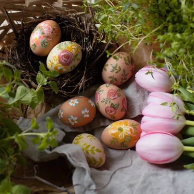 Colorful Easter eggs with floral patterns sit in a nest and on a cloth, next to pink tulips and green foliage.