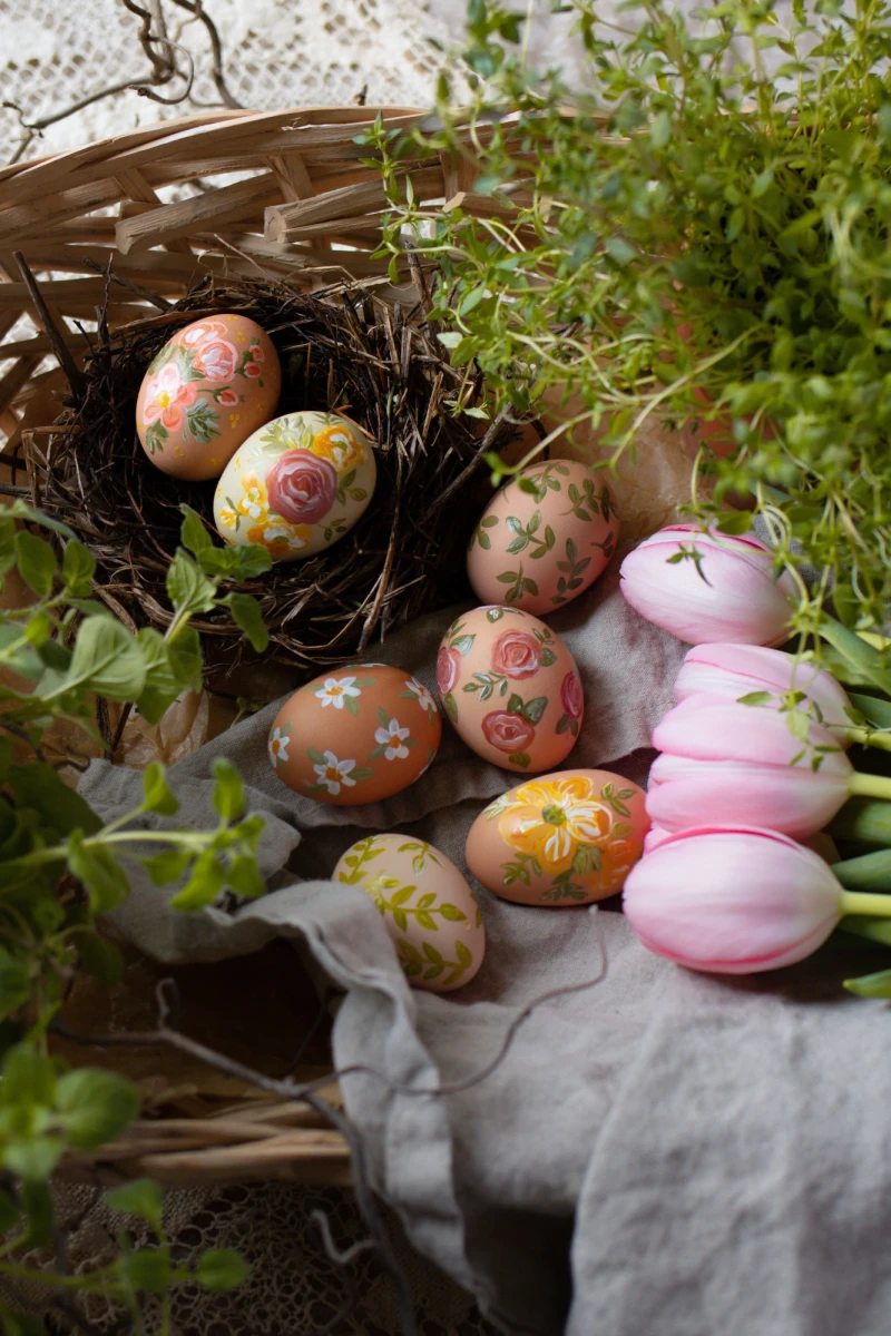 Colorful Easter eggs with floral patterns sit in a nest and on a cloth, next to pink tulips and green foliage.