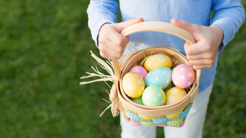 A person holding a pastel-colored basket filled with decorated Easter eggs, outdoors on green grass, ready for an egg hunt.