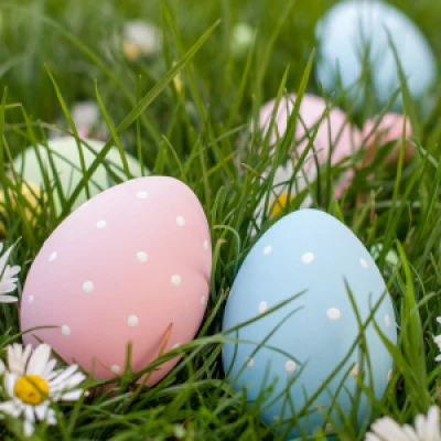 A festive spring décor: pastel hanging spirals, a bouquet of white daisies, colorfully decorated eggs in a basket, a green pitcher vase, and a decorative wreath on a red triangle, all on a bright windowsill.