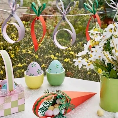 A festive spring décor: pastel hanging spirals, a bouquet of white daisies, colorfully decorated eggs in a basket, a green pitcher vase, and a decorative wreath on a red triangle, all on a bright windowsill.