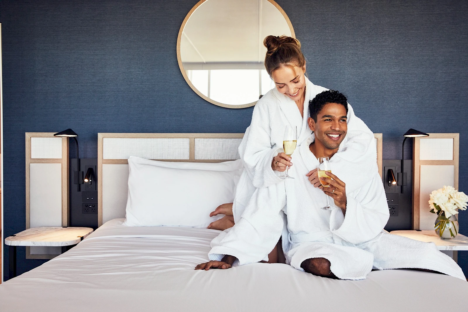 Two people in white robes sit on a hotel bed, smiling and toasting with champagne glasses. A round mirror and bedside lamps decorate the blue wall behind them.