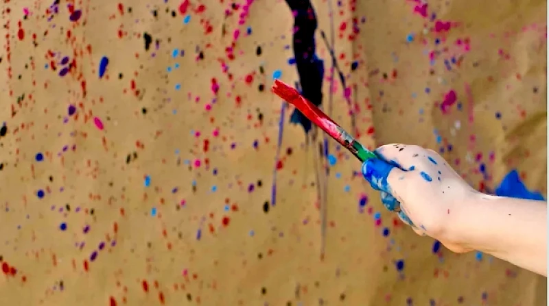 A close-up of a paint-splattered wall with a hand holding a red, blue-dyed paintbrush streaking color across the canvas, creating bold streaks.