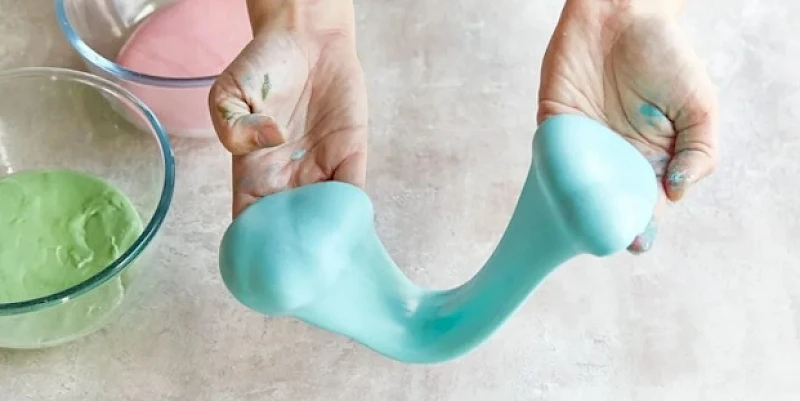 A person stretches turquoise slime into a curved shape, with bowls of green and pink slime nearby in the background.