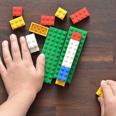 Two hands building with colorful LEGO bricks on a wooden surface, assembling a green-and-blue rectangular structure with scattered bricks around.