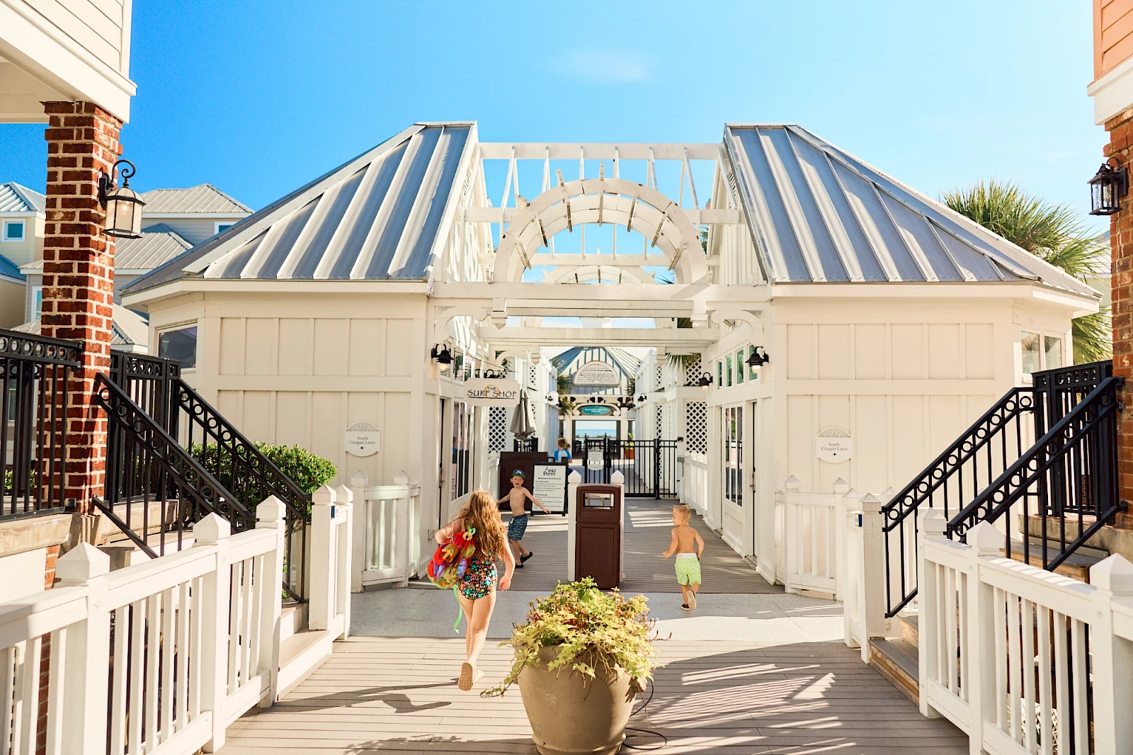 Outdoor white wooden walkway with glass-roofed arches, potted plants, and people walking under a bright blue sky, sunny seaside vibe.