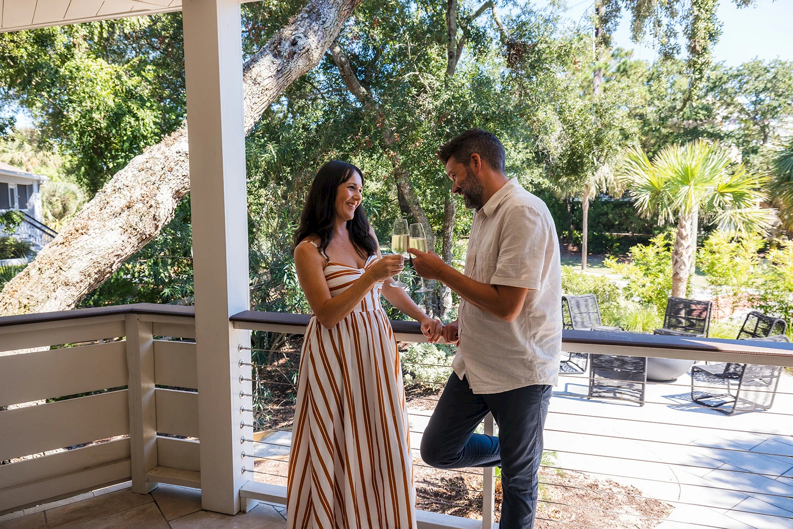 Two people on a deck share a toast with drinks, smiling as they stand under a tree-filled yard on a sunny day.