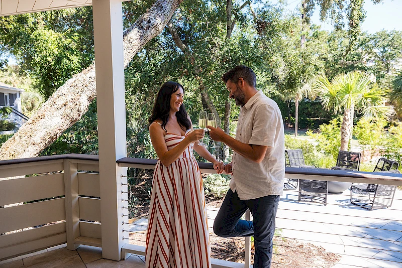 Two people on a deck share a toast with drinks, smiling as they stand under a tree-filled yard on a sunny day.