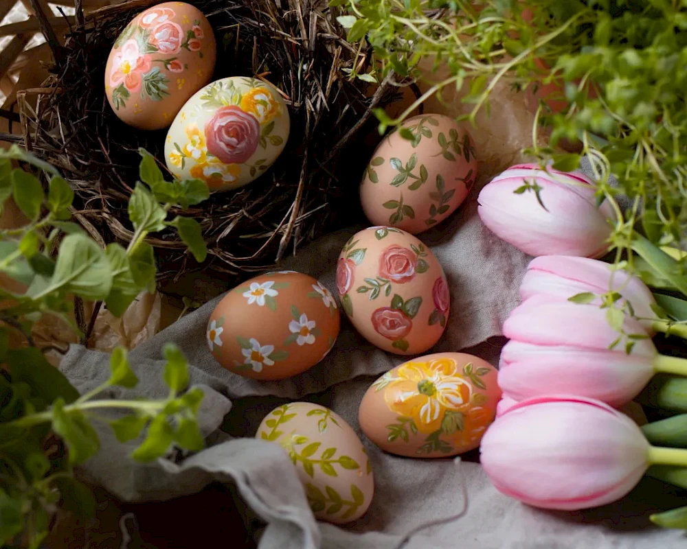 Colorful decorated Easter eggs in a woven basket with flowers and greenery, plus pink tulips and a soft fabric backdrop.