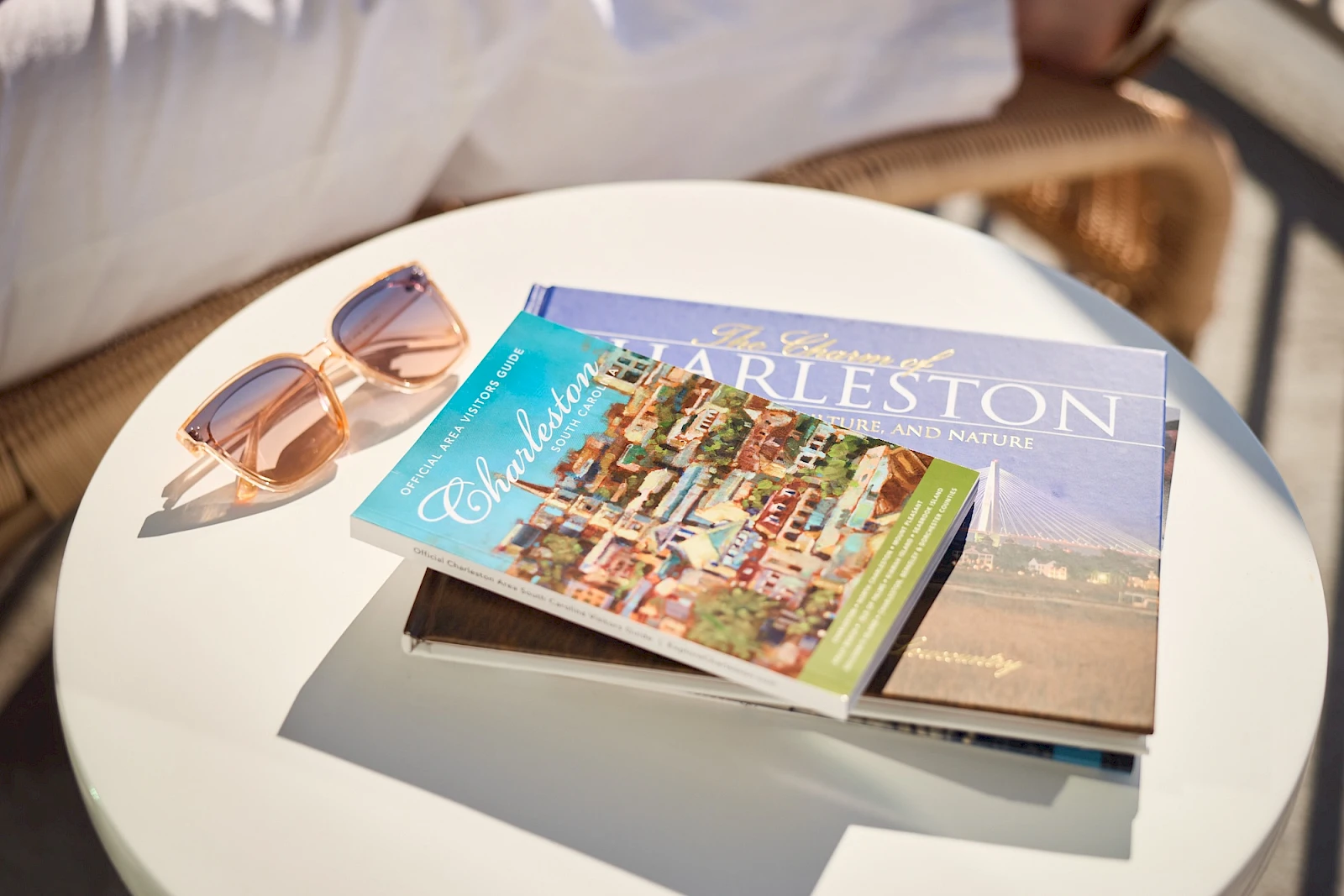 A small round table with two fashion/ travel magazines fanned open, a pair of sunglasses beside, and soft sunlight on a sunny outdoor setting.