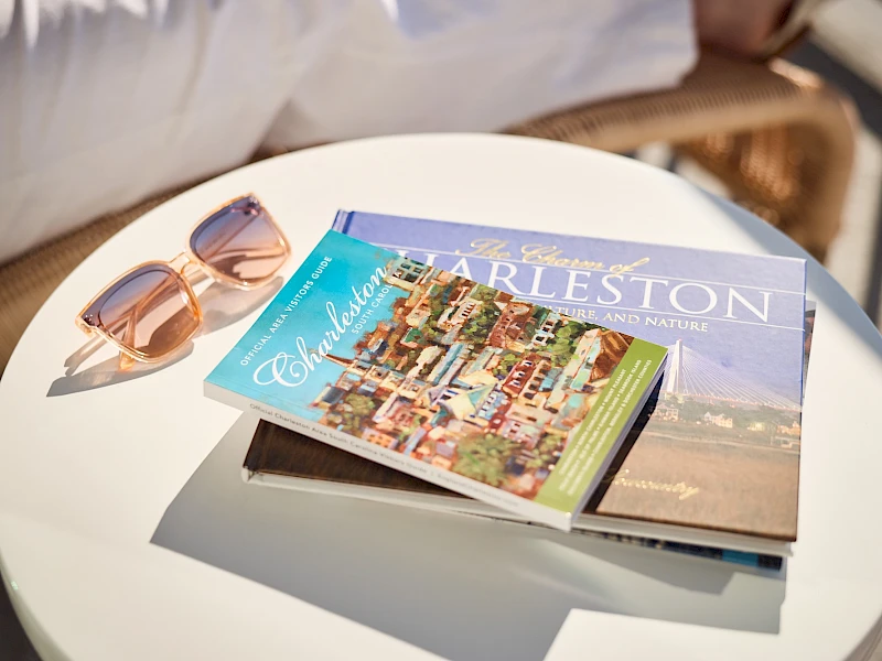 A small round table with two fashion/ travel magazines fanned open, a pair of sunglasses beside, and soft sunlight on a sunny outdoor setting.