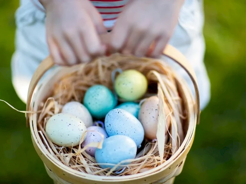 A hand-held basket of colorful speckled Easter eggs nestled in straw, with a green, grassy background.