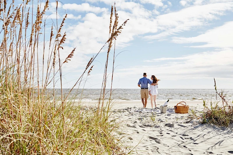 A couple walks hand in hand along a sandy beach with tall grasses, a picnic basket nearby, and the ocean under a partly cloudy sky, enjoying a sunny day.