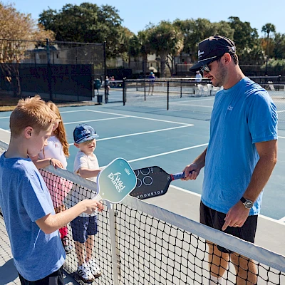 Two boys at a tennis lesson with a coach at a net, holding paddles on an outdoor court. The coach shakes hands with a boy while others wait, sunny day.