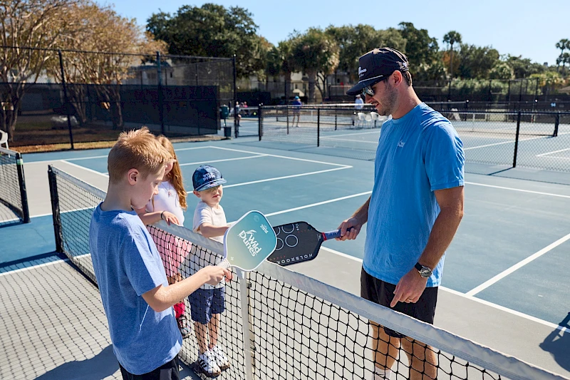 Two boys at a tennis lesson with a coach at a net, holding paddles on an outdoor court. The coach shakes hands with a boy while others wait, sunny day.