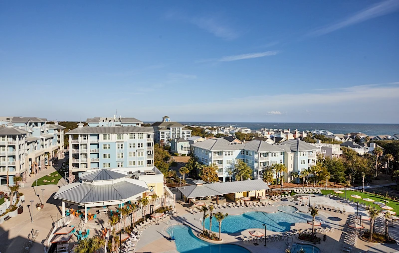 Coastal resort town with multiple low-rise buildings, a large central pool area, palm trees, and a bright blue sky over a calm sea.