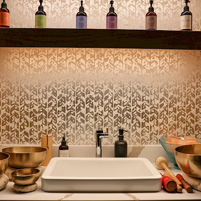 A kitchen or sink area with a modern white sink, brass bowls on the left, rolling pins on the right, bottles on a shelf above, and patterned tile backdrop, warmly lit.