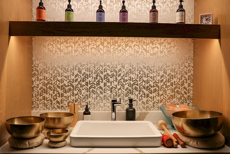 A kitchen or sink area with a modern white sink, brass bowls on the left, rolling pins on the right, bottles on a shelf above, and patterned tile backdrop, warmly lit.