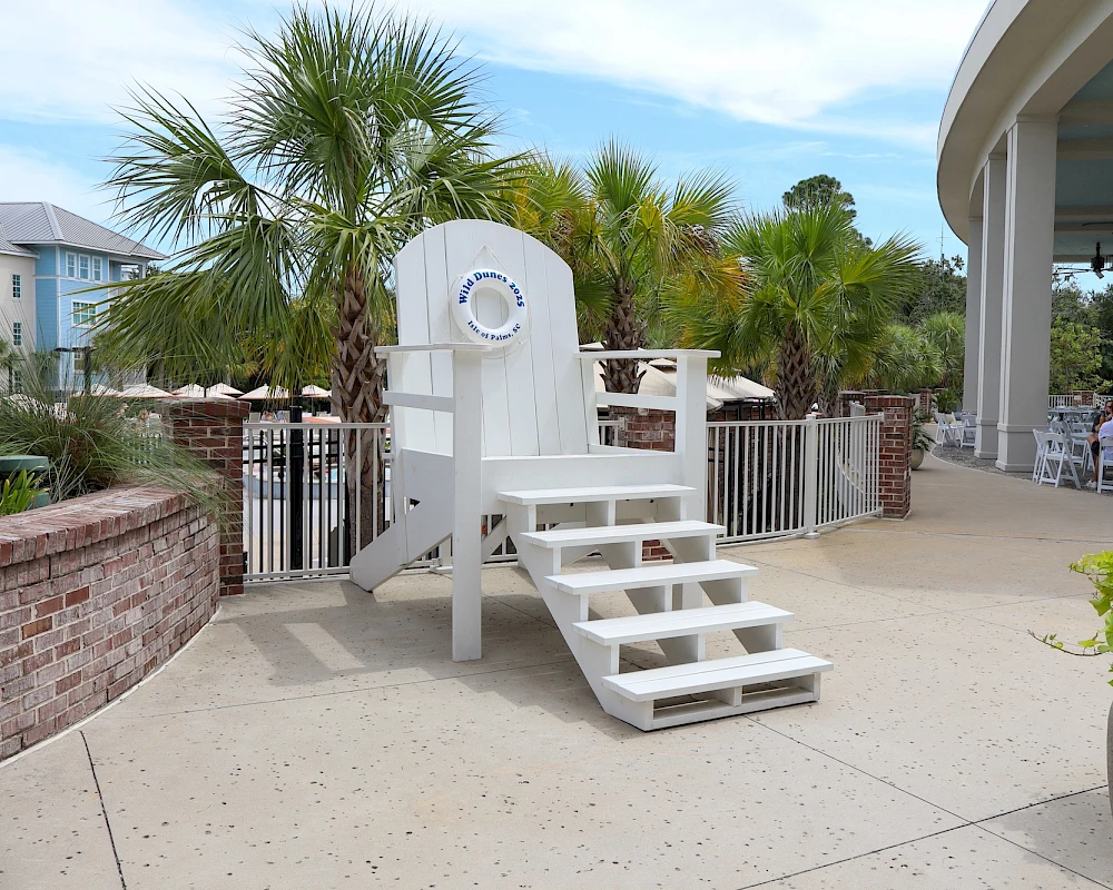 White lifeguard chair on a breezy patio by a curved building, palm trees, and potted plants along a sunny waterfront walkway.