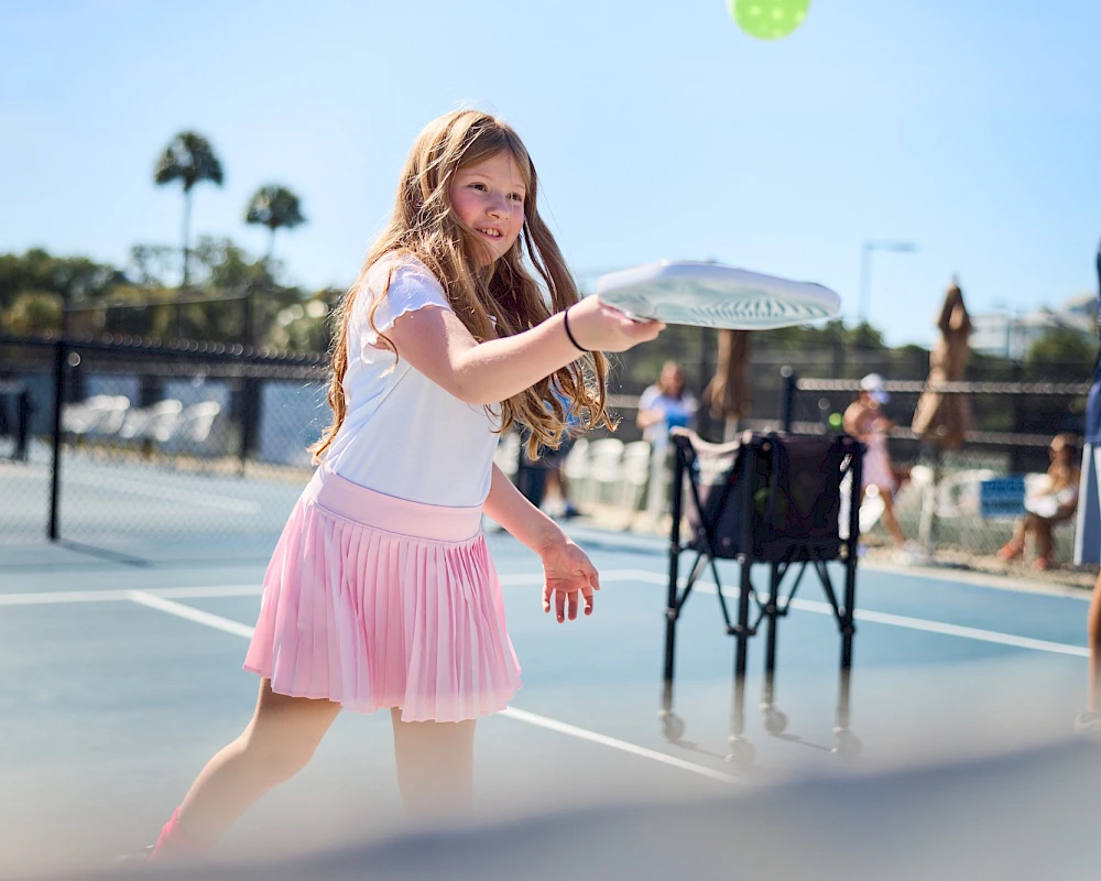 A girl in a pink skirt tosses a frisbee on a sunny tennis court; others watch and play in the background.