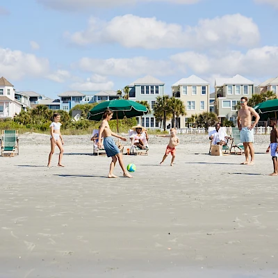 People play on a sunny beach: kids kick a ball, adults chat, beach chairs and green umbrellas line the sandy shore, pastel houses in the background.