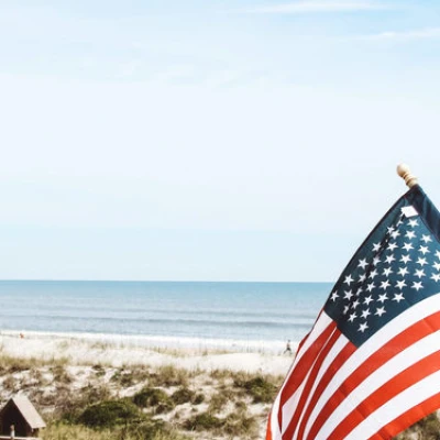 American flag waving on a sunny beach with dunes and calm sea in the background.
