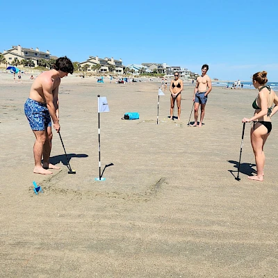 Kids playing a beach game with sticks and cups in the sand, a sunny day, and onlookers nearby, all near the shoreline.
