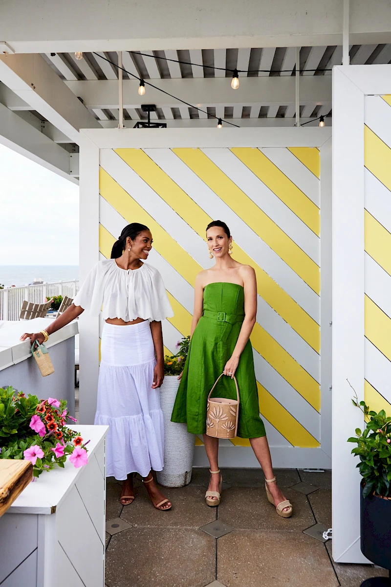 Two women pose on a balcony-like set with yellow diagonal stripes, one in a white off-shoulder top and lilac skirt, the other in a green dress.