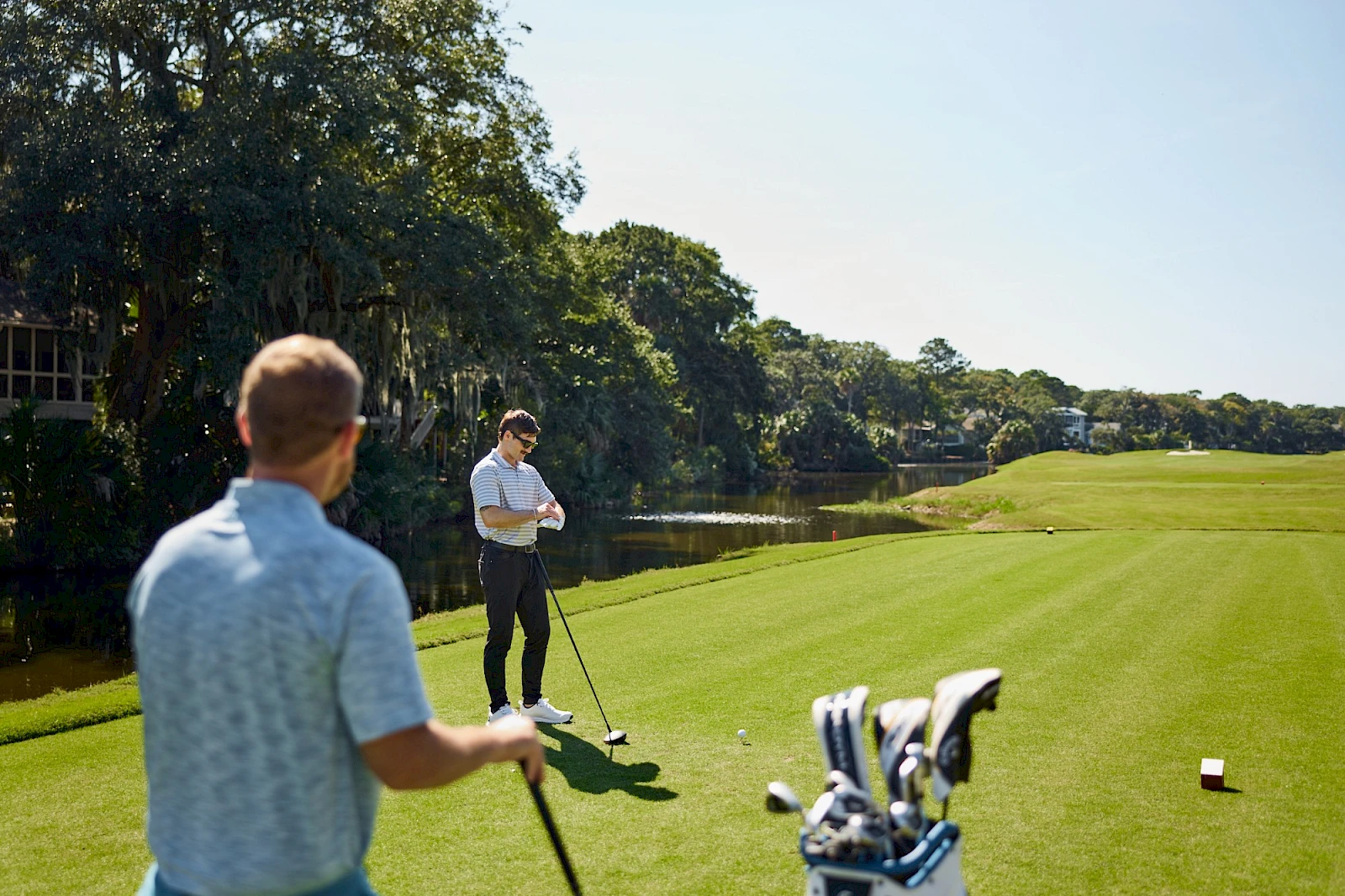 Two men playing golf on a sunny course, one swinging while the other watches near a set of golf bags and a lake in the background.