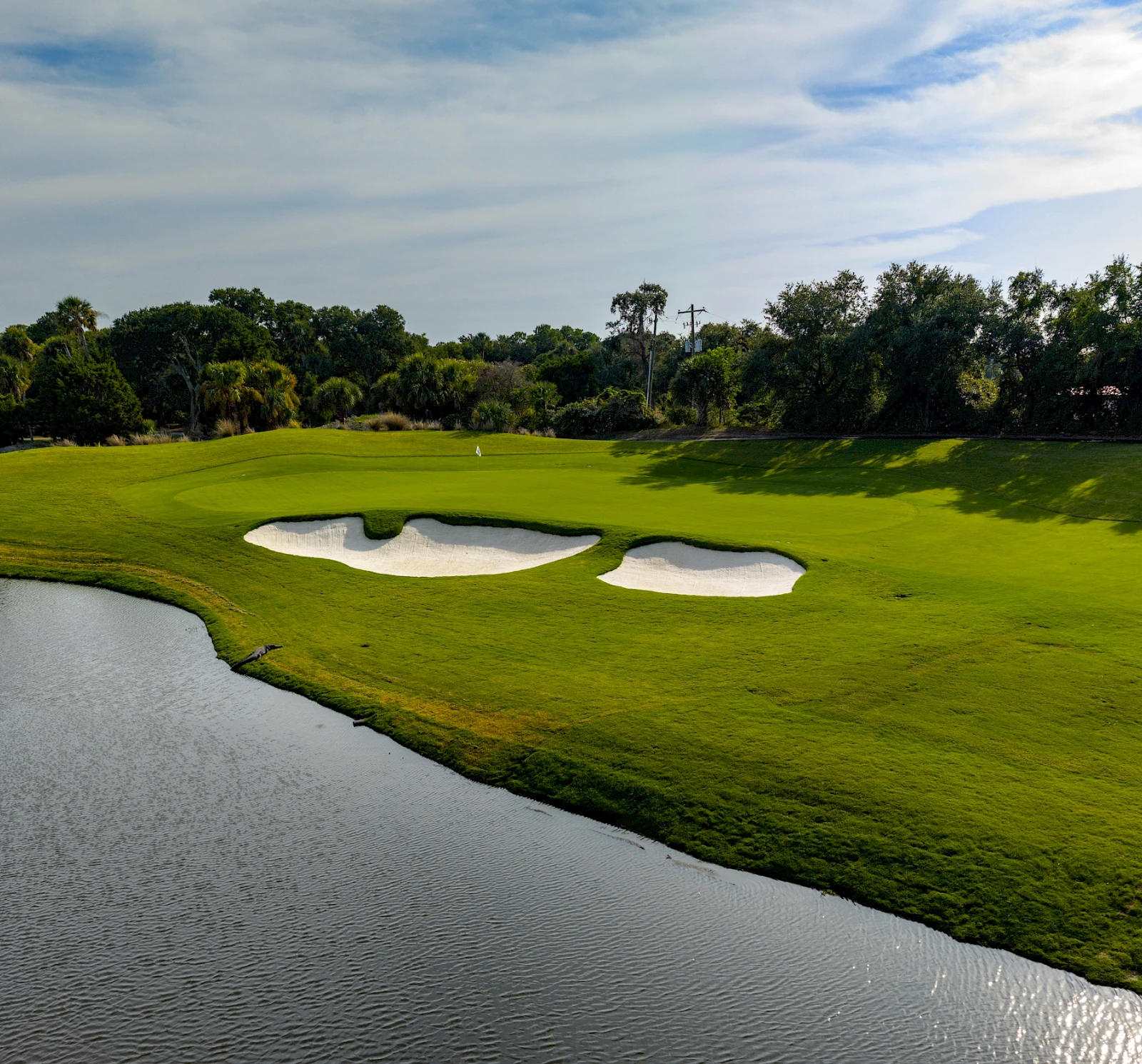 A lush golf course with green fairways, sand bunkers, and a water hazard surrounding the greens, under a partly cloudy sky.