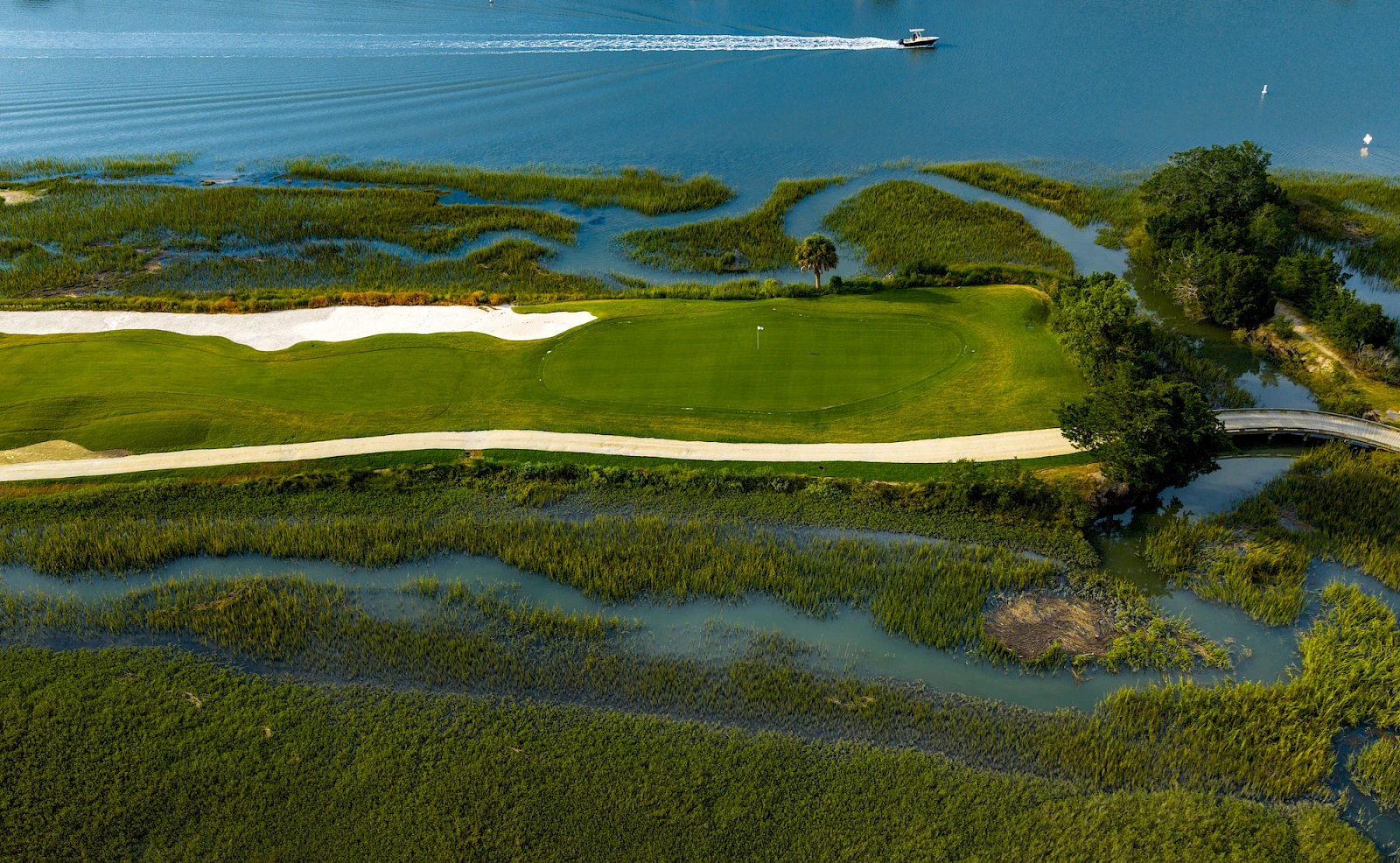 An aerial view of a golf course fairway and greens bordered by winding water channels and marshy inlets, with a boat visible in the water.