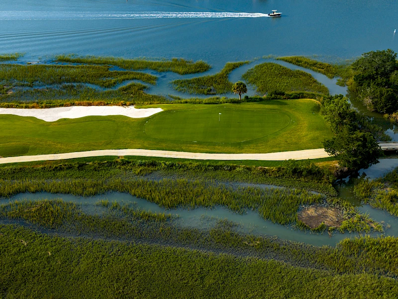 An aerial view of a golf course fairway and greens bordered by winding water channels and marshy inlets, with a boat visible in the water.