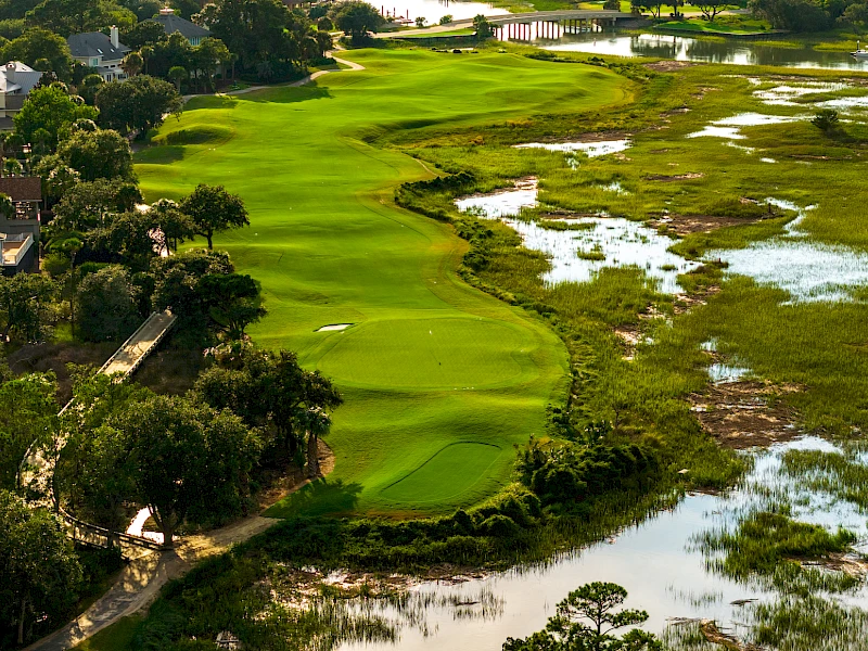 An aerial view of a lush golf course winding through green fairways, bordered by trees and water, with residential areas along the left edge.
