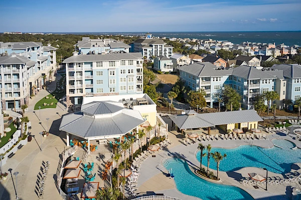 Aerial view of a coastal resort with multiple white buildings, a large pool area with palm trees, lounge chairs, and a central gazebo.