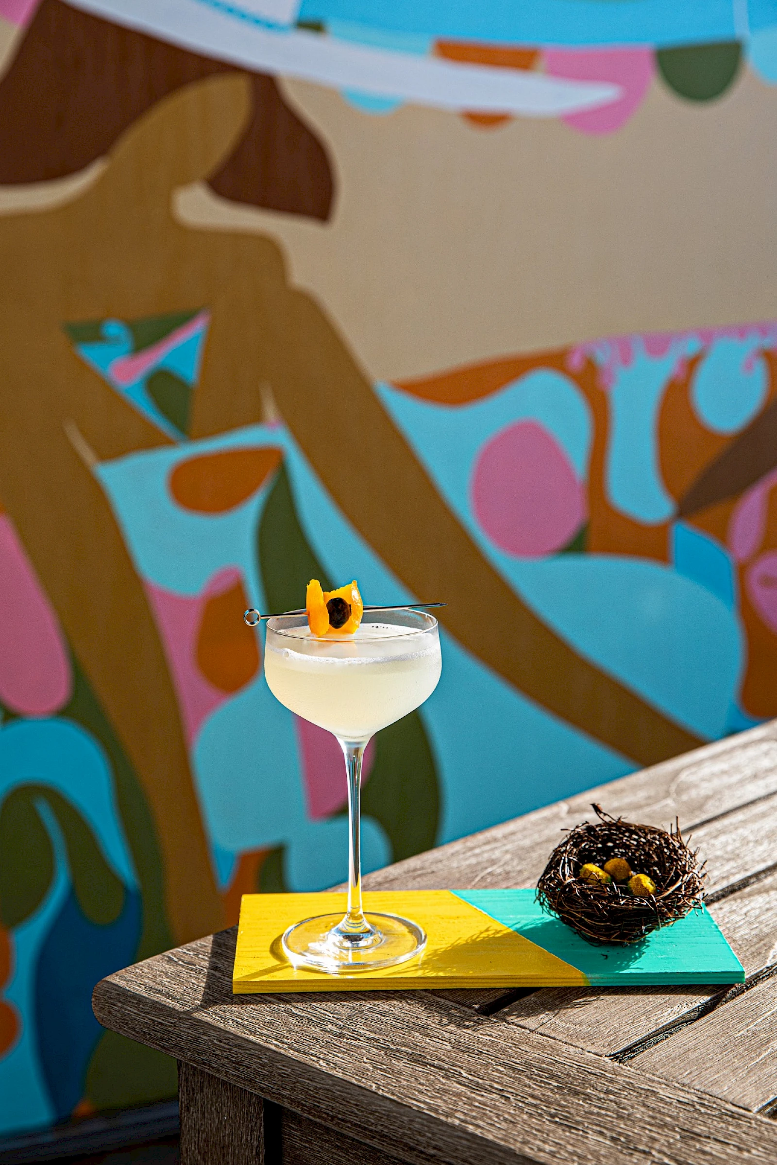 A cocktail on a stemmed glass with an orange garnish sits on a yellow napkin atop a teal book on a sunlit wooden surface; a spiky plant lies nearby. The colorful mural in the background adds a vibrant, abstract vibe.