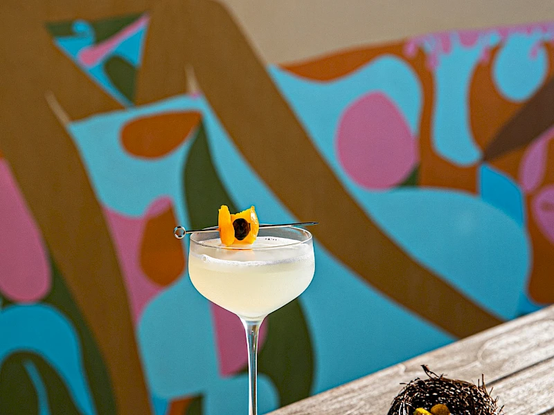 A cocktail on a stemmed glass with an orange garnish sits on a yellow napkin atop a teal book on a sunlit wooden surface; a spiky plant lies nearby. The colorful mural in the background adds a vibrant, abstract vibe.