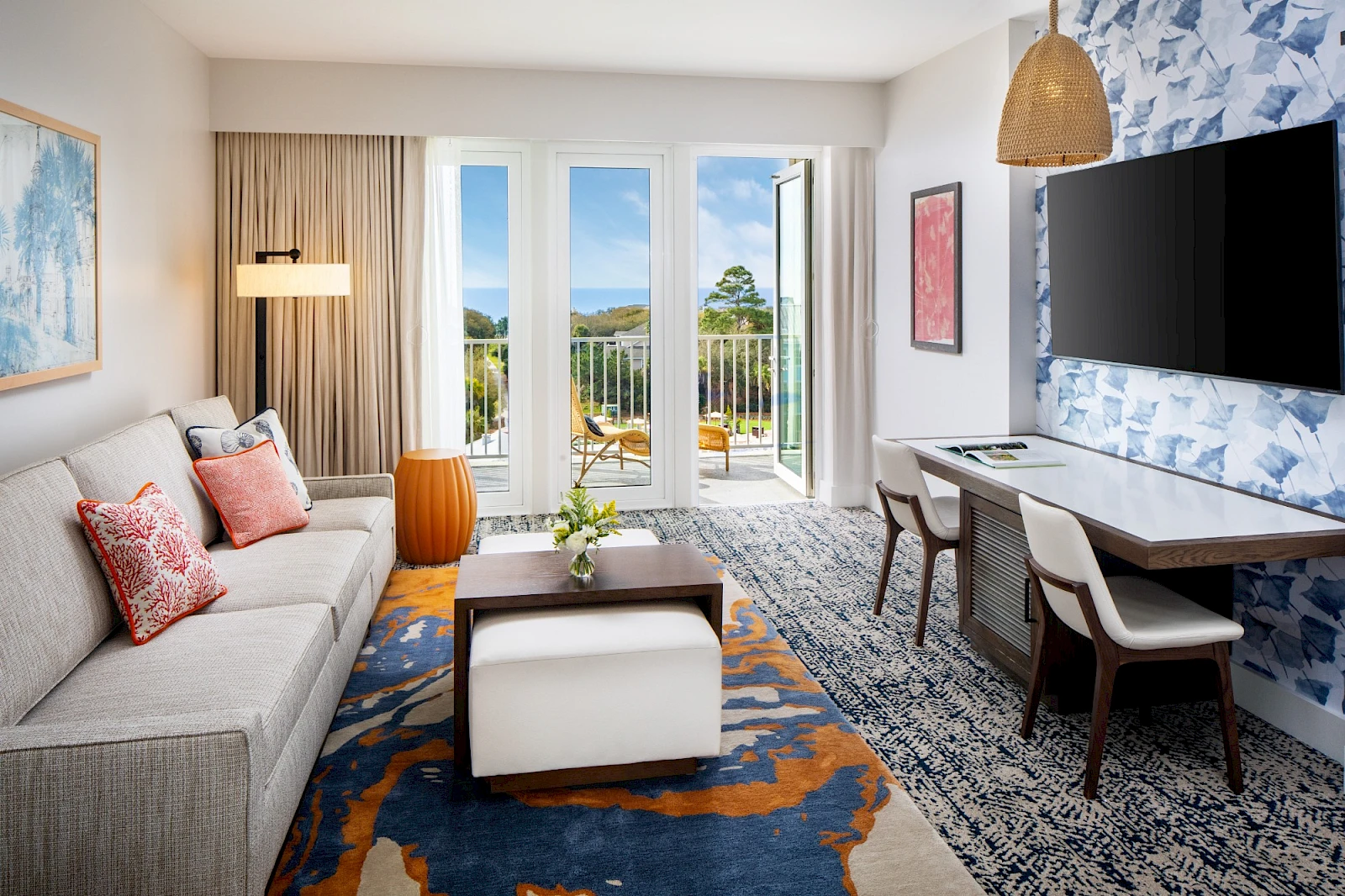 Stylish living room with a gray sofa, colorful pillows, coffee table, patterned rug, and a wall-mounted TV; glass doors open to a balcony with greenery.