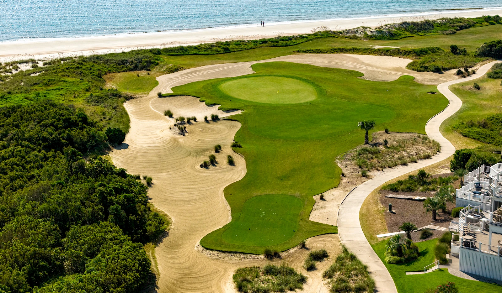 An aerial view of a coastal golf course: green fairways, sand traps, dunes, and ocean backdrop, with a winding path and nearby buildings.