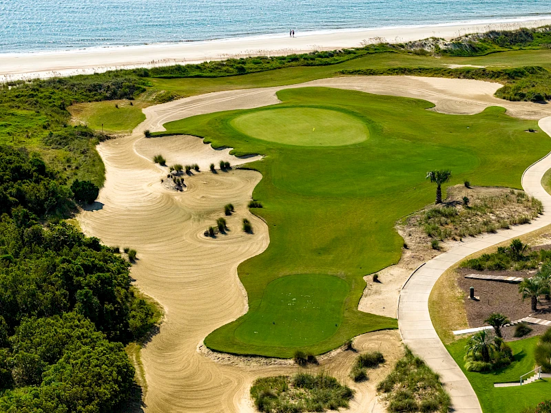 An aerial view of a coastal golf course: green fairways, sand traps, dunes, and ocean backdrop, with a winding path and nearby buildings.