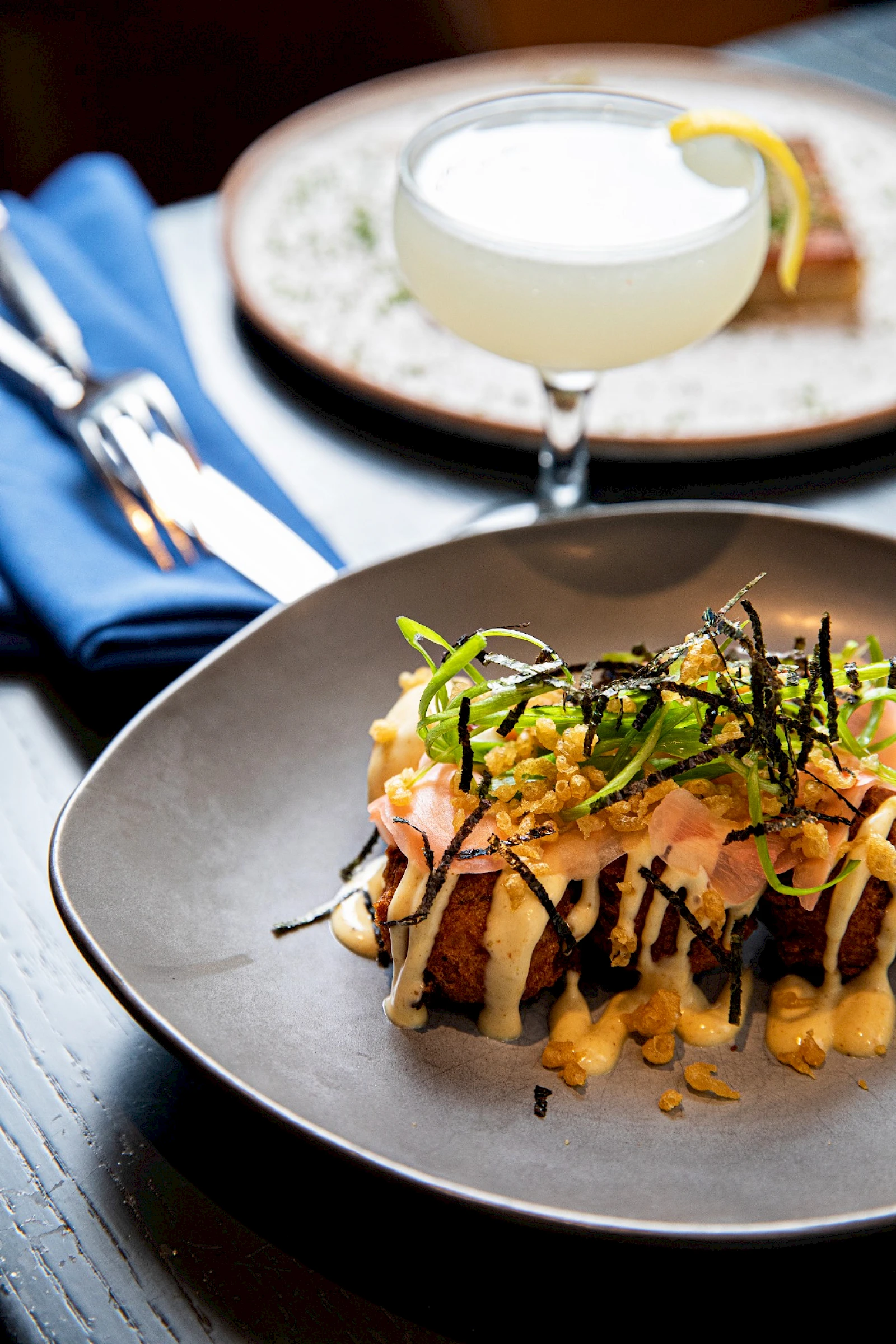A plated gourmet dish topped with crispy onions and greens, with a cocktail in the background and a slice of lime garnish.