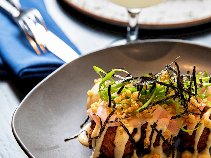 A plated gourmet dish topped with crispy onions and greens, with a cocktail in the background and a slice of lime garnish.