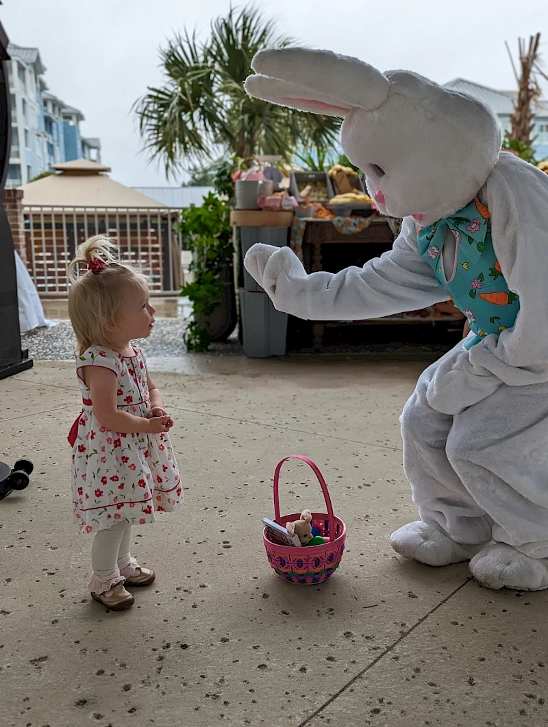 A child in a floral dress meets a person in a bunny costume offering a hello high-five near a pink basket, outdoors on a sunny day.