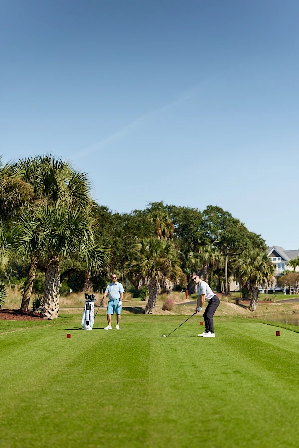Three people at a sunny golf course tee off: a child with a parent watching, and a player about to swing on well-manicured grass.