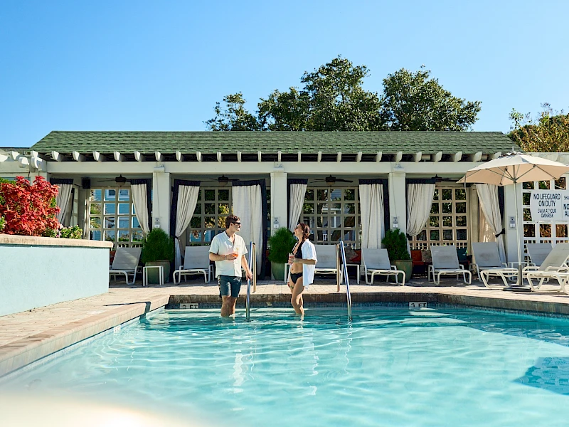 Two people walk into a sunny pool, lounge chairs and an umbrella-set terrace behind, a white-walled clubhouse with green roof in the background.