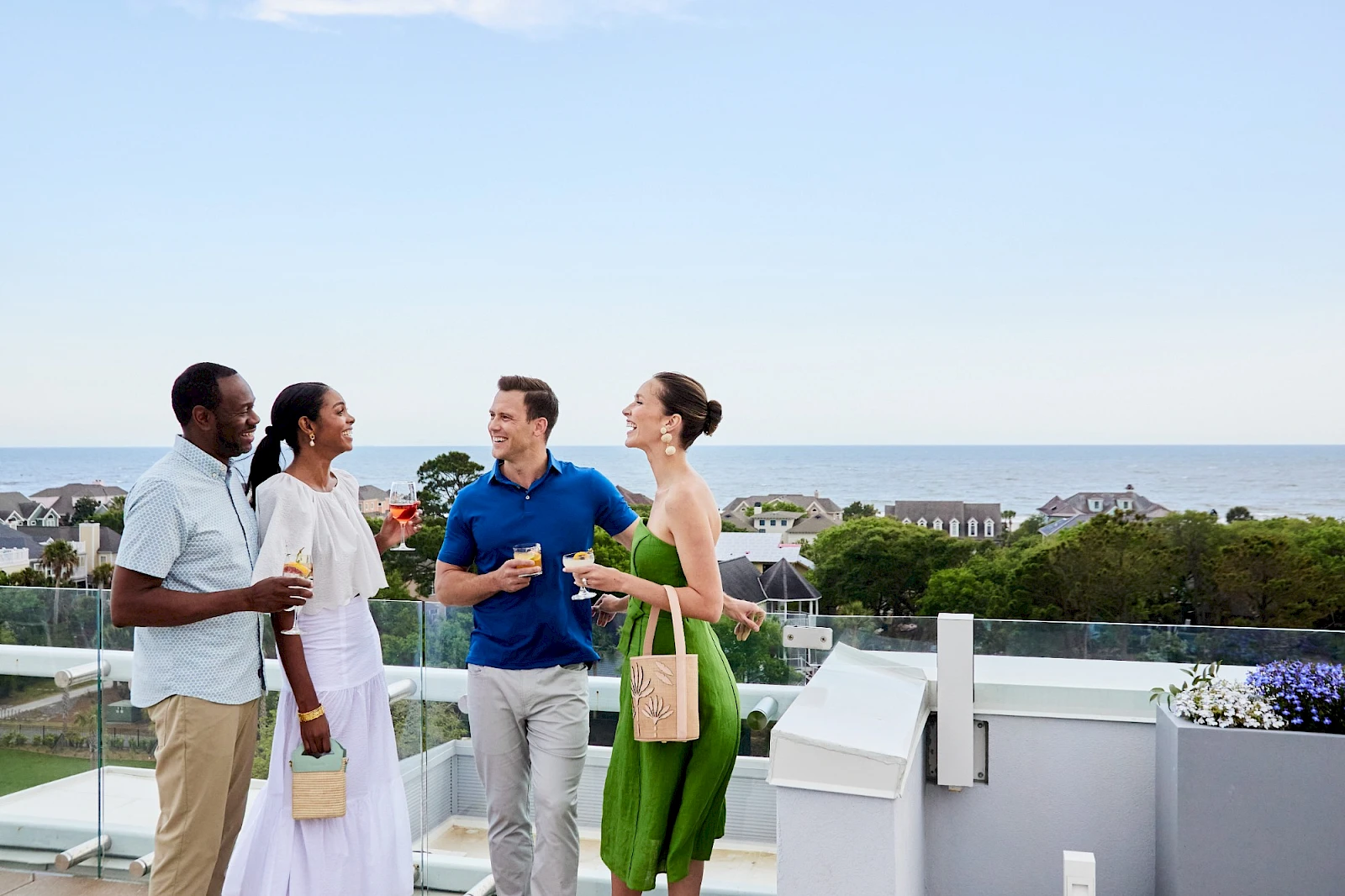 Four friends chat on a balcony overlooking the ocean, sharing drinks and smiles at a sunny rooftop gathering.