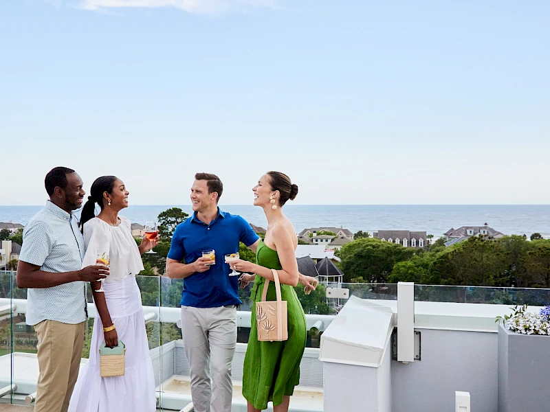 Four friends chat on a balcony overlooking the ocean, sharing drinks and smiles at a sunny rooftop gathering.