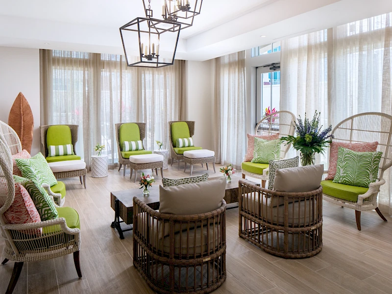 A bright, airy living room with rattan chairs, lime-green cushions, pale curtains, and a circular seating arrangement around a small table. Ends with a period.