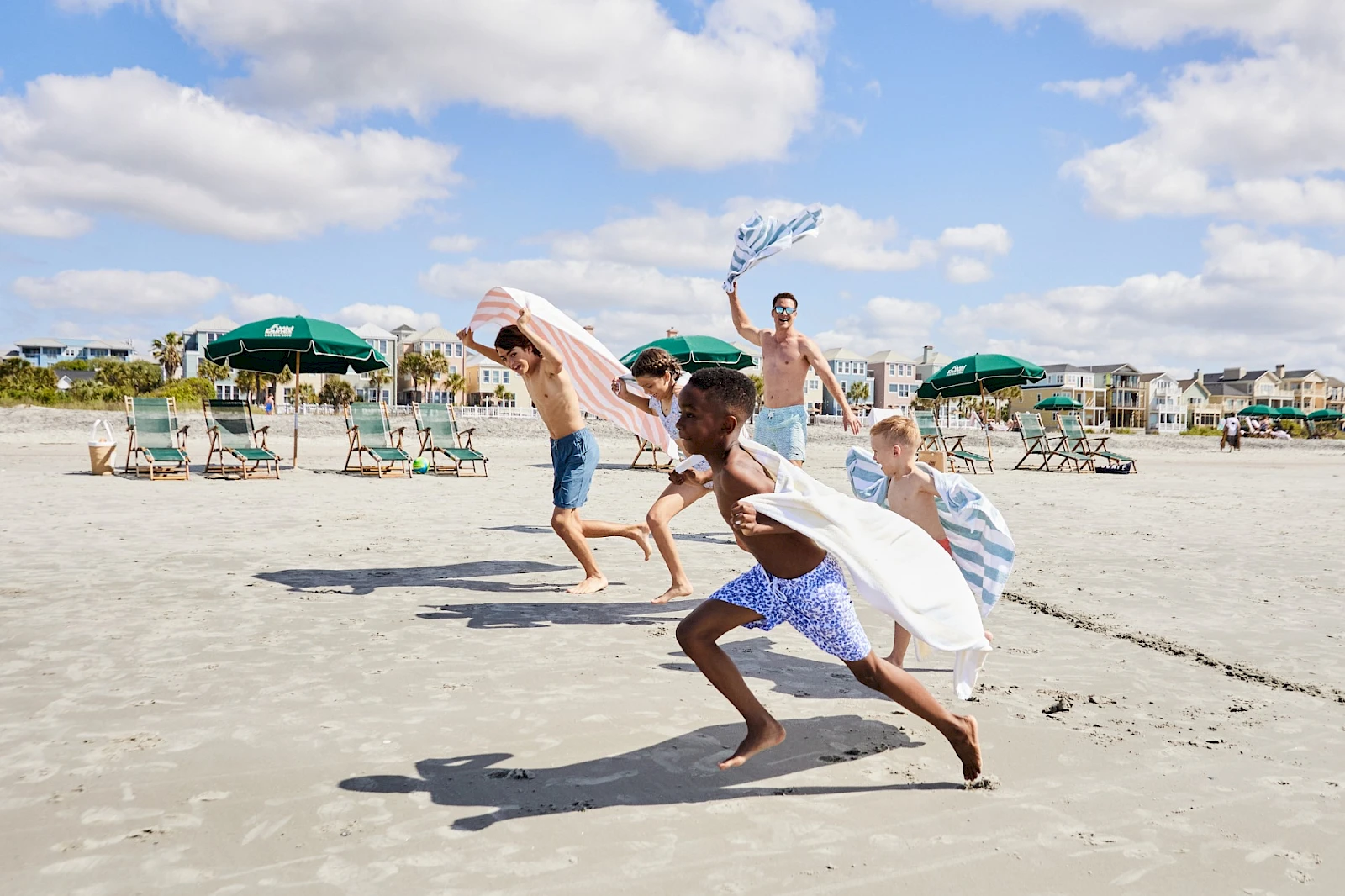 Four friends race along the sunny beach, towels and a sheet fluttering like sails as they sprint past green beach umbrellas on warm sand.