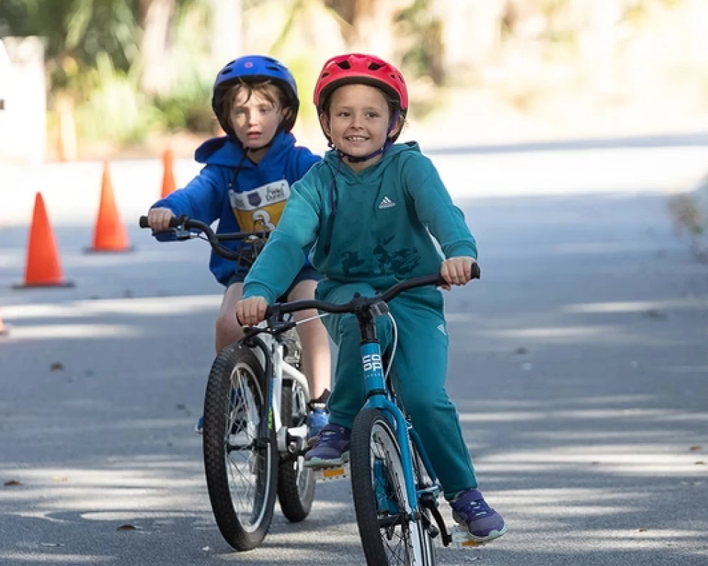 Two kids ride balance bikes on a sunny path, wearing helmets, with orange cones in the background, enjoying a cheerful outdoor ride.