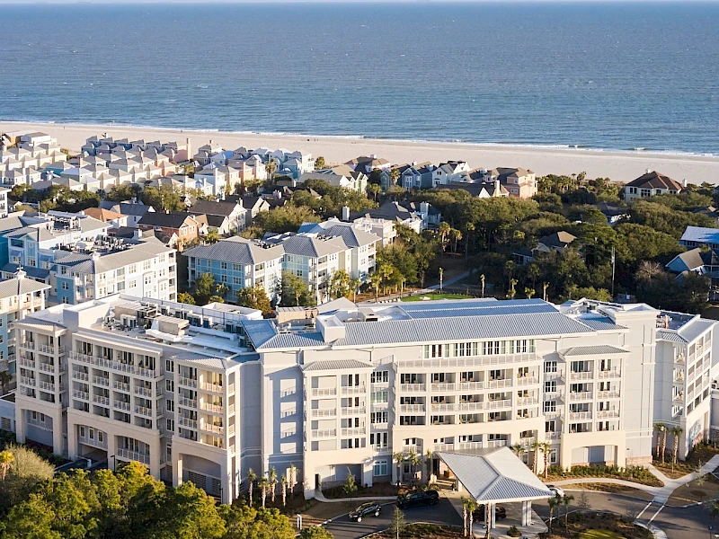 A coastal coastal town with a large white apartment complex, sandy beach, and ocean beyond, surrounded by trees and greenery.